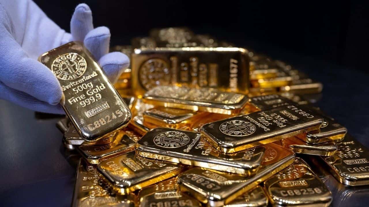 دست یک نفر با دستکش سفید در حال نشان دادن یک قطعه طلای آبشده از بین تعداد زیادی قطعه دیگر
